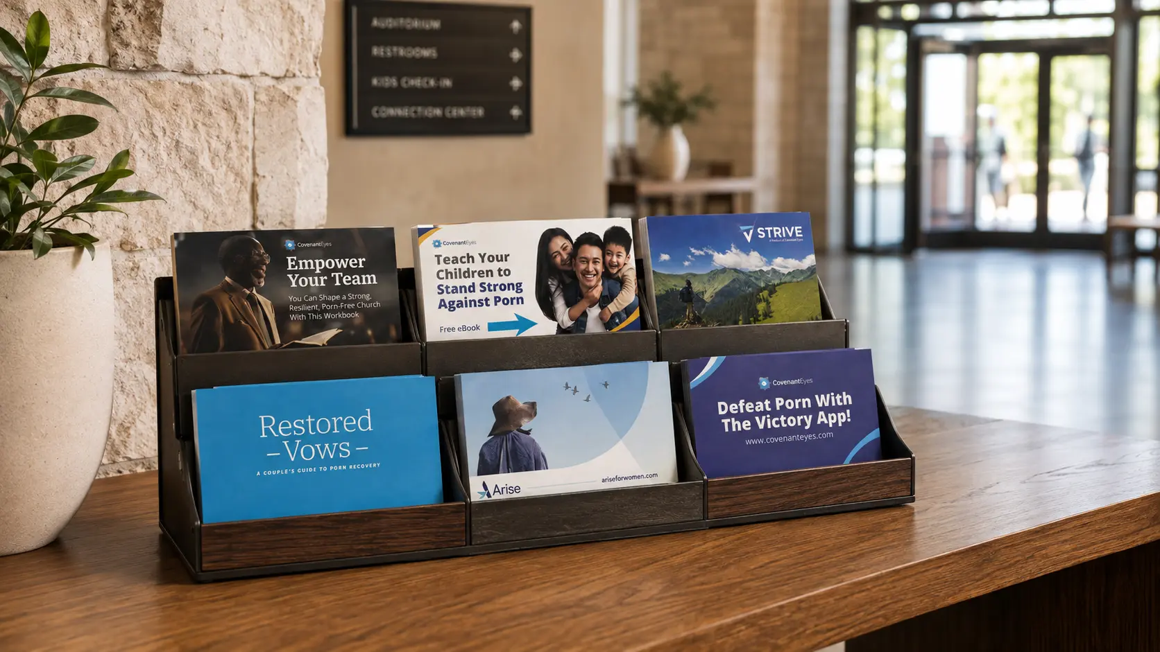 A wooden six-card display on a counter in a church vestibule, holding the leader, parents, men&rsquo;s, couples, women&rsquo;s and Victory App cards, with the entrance doors behind.